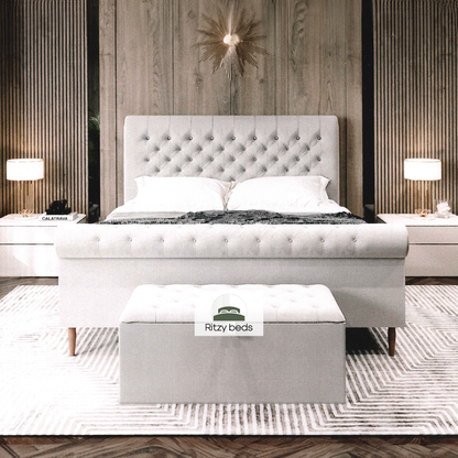 Gray upholstered handmade bed with buttoned tufted scrool headboard and footboard in a bedroom setting, featuring 'Ritzy Beds' branding.