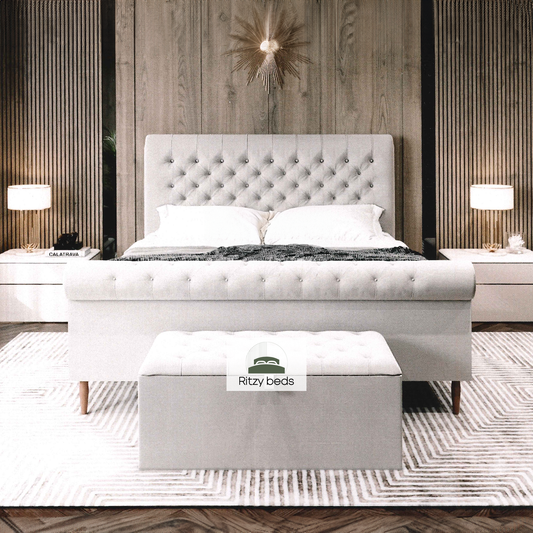 Gray upholstered handmade bed with buttoned tufted scrool headboard and footboard in a bedroom setting, featuring 'Ritzy Beds' branding.